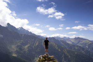 Hiker standing on a rocky summit, looking out over a vast mountain valley under a blue sky