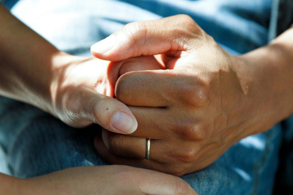 Close-up of one person's hands gently clasping another's, a thin ring on one finger, resting on denim