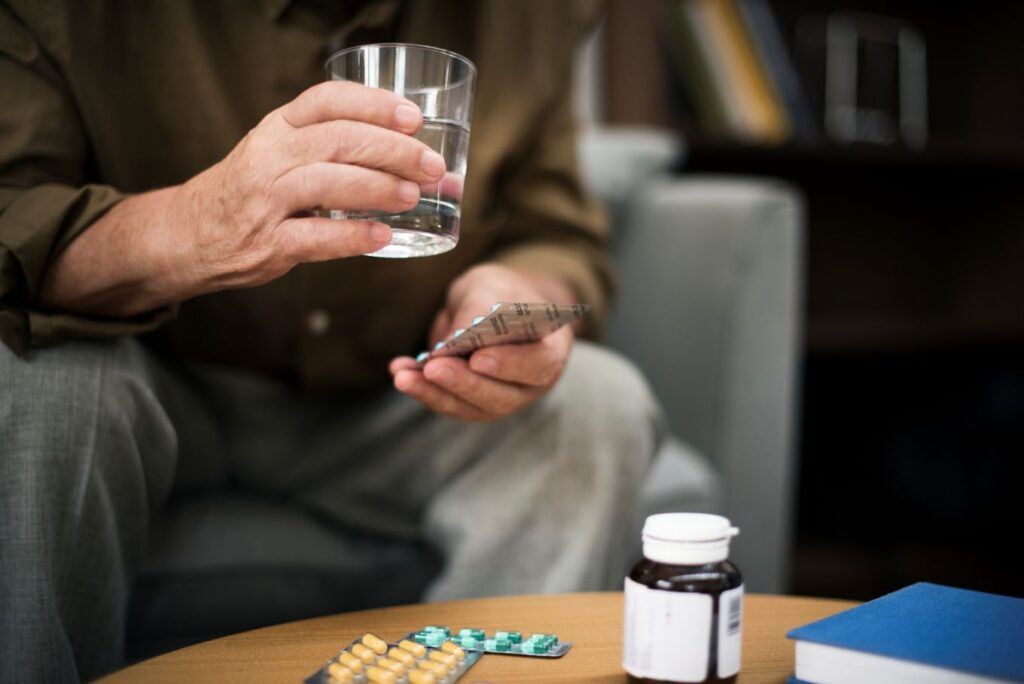 Older man seated in armchair holding a glass of water and a blister pack of tablets, with pill bottle nearby