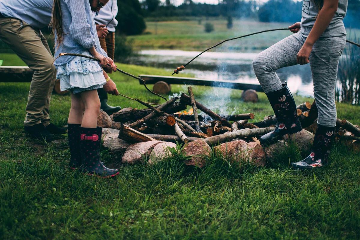 Family in wellies roasting food on sticks over a campfire beside a lake