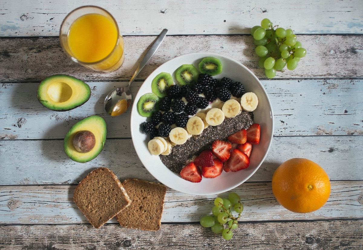 Bowl of yoghurt topped with blackberries, kiwi, banana, chia seeds and strawberries, surrounded by avocado, wholemeal bread, grapes, orange and juice