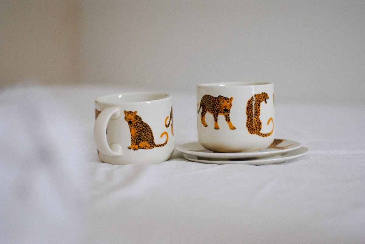 Two cream ceramic mugs decorated with orange leopards, one on a saucer, on white bedding