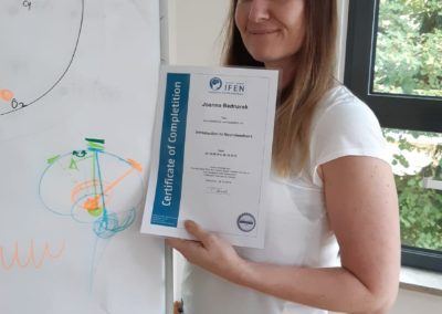 Smiling woman holding an IFEN Certificate of Completion beside a flipchart with hand-drawn diagrams