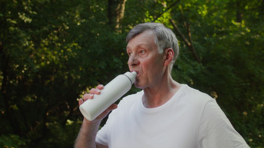 Older man with grey hair drinking from a white bottle outdoors, surrounded by green woodland