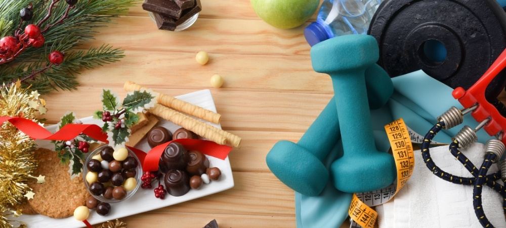 Christmas chocolates and biscuits beside teal dumbbells, a tape measure and resistance bands on a wooden table