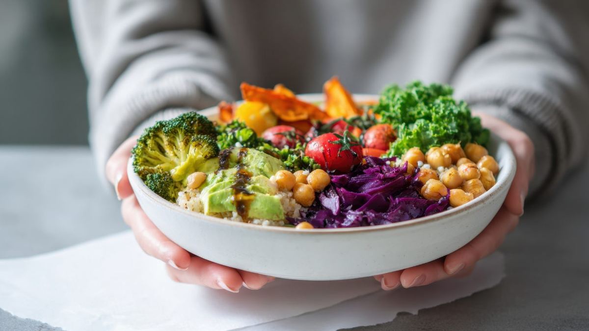 Hands holding a colourful bowl of broccoli, avocado, chickpeas, red cabbage, kale, tomatoes and quinoa