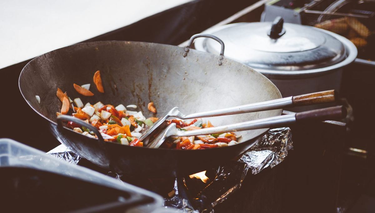 Chopped carrots, onions and peppers stir-frying in a large wok over a flame, with two spatulas