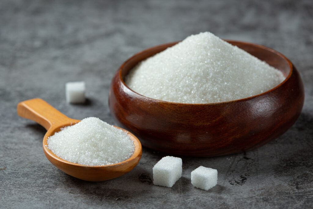 Granulated sugar heaped in a dark wooden bowl and on a wooden spoon, with sugar cubes nearby