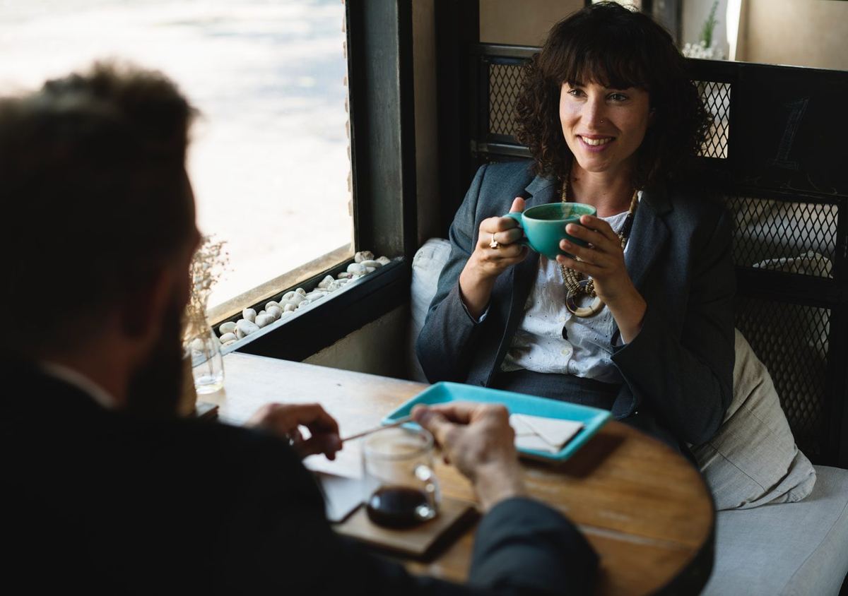 Smiling woman holding a teal cup across a café table from a bearded man stirring a drink