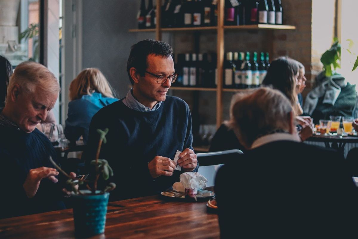 Man in glasses sitting at a café table with older companions, wine bottles on shelves behind them